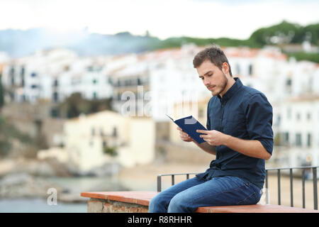 Homme sérieux lire un livre papier assis sur une corniche dans une ville côtière Banque D'Images