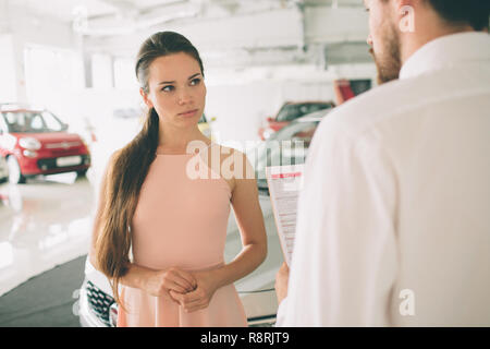 Vendeur de voiture sympathique parlant à une jeune femme et montrant une nouvelle voiture à l'intérieur de la signature de contrat d'exposition. Banque D'Images