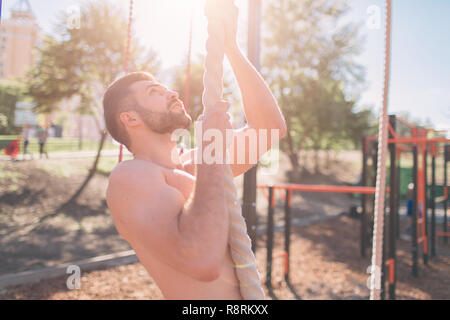 Homme barbu avec des cheveux noirs escalade corde épaisse au cours d'exercices en cross-fit avec la formation. Mains musculaire très fort. Banque D'Images