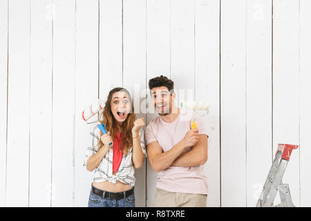 Photo de couple homme et femme joyeuse peinture mur blanc et faire de la rénovation à l'aide de rouleaux à peinture Banque D'Images