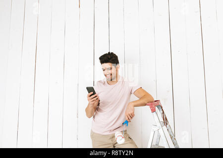 Photo de jeune homme 20s à l'aide téléphone portable en peinture mur blanc avec rouleau à peindre et faire de la rénovation Banque D'Images