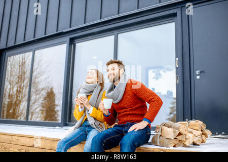 Quel beau couple de jeunes vêtus de chandails colorés assis profiter de la nature ainsi que des boissons chaudes sur la terrasse de la maison moderne dans les montagnes Banque D'Images