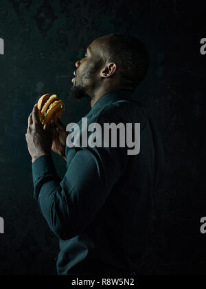 La young african american man eating hamburger et à l'écart sur fond de studio noir Banque D'Images