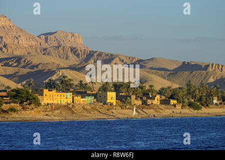 Vue d'un petit village typique en bord de rivière, sur les rives du Nil, en Haute-Égypte, avec des palmiers contre un désert aride colline historique Banque D'Images