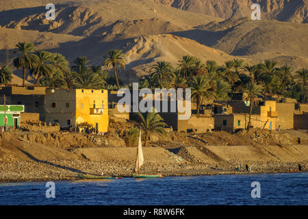 Vue d'un petit village typique en bord de rivière, sur les rives du Nil, en Haute-Égypte, avec des palmiers contre un désert aride colline historique Banque D'Images