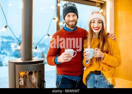 Jeune couple vêtus de chandails lumineux et de chapeaux de standing ainsi que des boissons chaudes près de la cheminée dans la maison moderne durnig l'heure d'hiver Banque D'Images