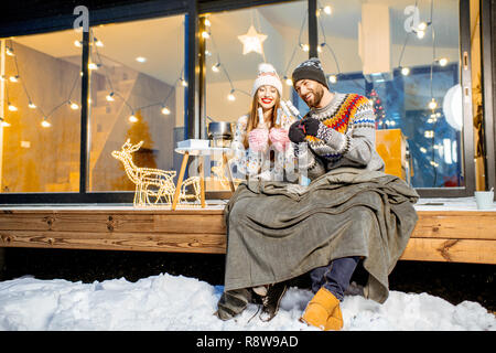 Jeune couple de préchauffage avec plaid et marshmallow assis sur la terrasse de la maison moderne dans les montagnes durnig les vacances d'hiver Banque D'Images