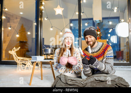 Jeune couple de préchauffage avec plaid et marshmallow assis sur la terrasse de la maison moderne dans les montagnes durnig les vacances d'hiver Banque D'Images