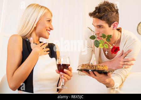 Un jeune homme avec une rose dans sa bouche, gâteau d'anniversaire de belle fille blonde, fille agréablement surpris en le regardant avec un sourire sur son visage. Banque D'Images