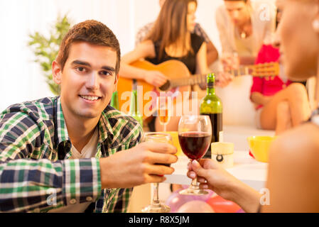 Un jeune homme avec un verre de vin à la maison partie toast avec une fille, et à l'arrière-plan vous pouvez voir un groupe de jeunes gens rassemblés autour de la g Banque D'Images