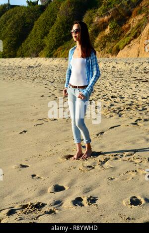 Jeune femme aux pieds nus, portant des lunettes et un jean, est debout sur la plage de Laguna Beach, en Californie. Banque D'Images