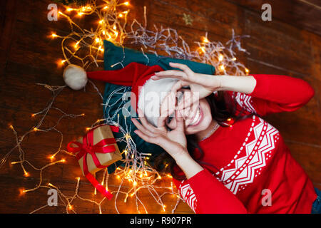 Close-up portrait of woman in Santa hat enveloppé dans les lumières de Noël s'amusant et lying on floor Banque D'Images