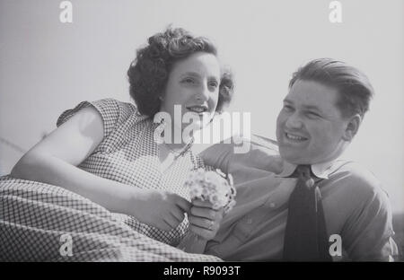 Années 1950, historique, un jeune couple se trouvant près de chaque ensemble en dehors, avec la jeune fille tenant un bouquet de fleurs Daisy, England, UK. Banque D'Images