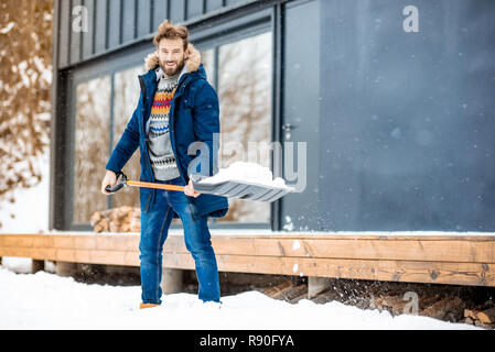 Bel homme dans des vêtements d'hiver neige nettoyage avec une pelle près de la maison moderne dans les montagnes Banque D'Images