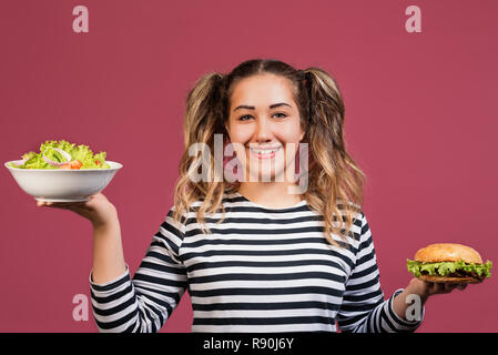 Cool girl with ponytails et t-shirt à rayures dans le choix entre une salade fraîche saine et malsaine sur fond rose coloré hamburger Banque D'Images