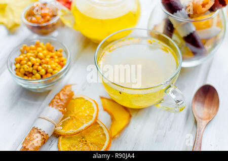 L'argousier thé dans la théière et tasse en verre et de nombreux fruits divers et de pastilles, plaquettes sur le petit déjeuner sur la table en bois blanc, lumière naturelle, l'accent selektive Banque D'Images