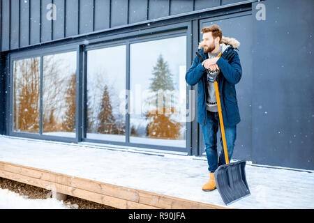 Portrait d'un homme dans des vêtements d'hiver debout avec pelle à neige sur la terrasse de l'immeuble dans les montagnes Banque D'Images