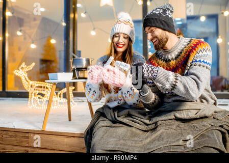 Jeune couple de préchauffage avec plaid et marshmallow assis sur la terrasse de la maison moderne dans les montagnes durnig les vacances d'hiver Banque D'Images