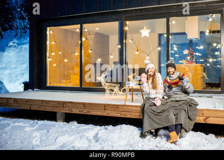 Jeune couple de préchauffage avec plaid et marshmallow assis sur la terrasse de la maison moderne dans les montagnes durnig les vacances d'hiver Banque D'Images