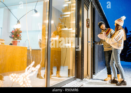 Jeune couple venant dans la magnifique maison décorée durnig les vacances d'hiver dans les montagnes enneigées Banque D'Images