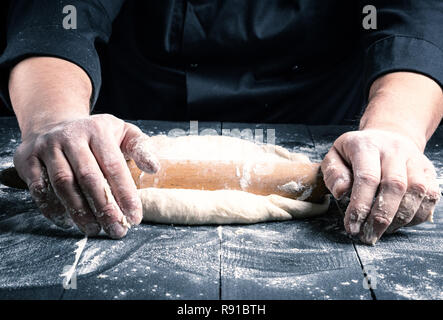 Chef dans une tunique noire roule une pâte à pizza ronde sur un tableau noir avec un rouleau à pâtisserie en bois Banque D'Images