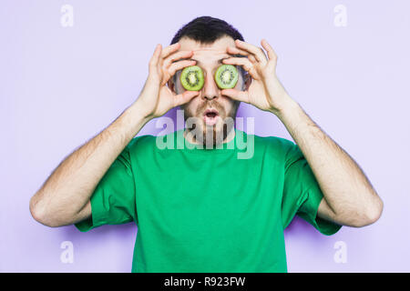 Jeune homme barbu tenant tranches de kiwi vert devant ses yeux, surpris et choqué. Fond mauve, copiez l'espace. Banque D'Images