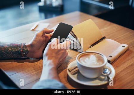 Les mains de l'homme en chemise en jean glisser avec le doigt sur l'écran de son téléphone intelligent, près de tasse à café sur table en bois Banque D'Images