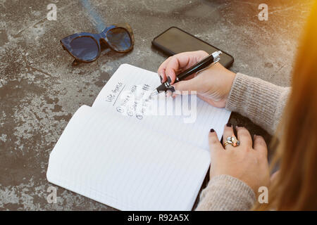 Jeune femme écrit sa vie avec des objectifs du papier et un stylo. Lunettes de soleil et téléphone cellulaire sont sur la table Banque D'Images