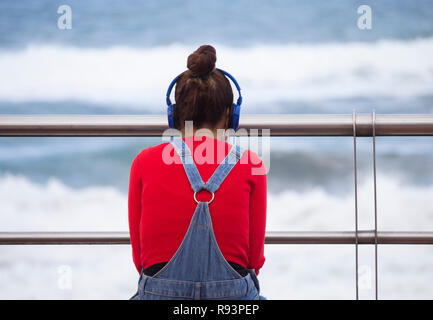 Vue arrière du jeune femme portant des écouteurs assis sur son fauteuil avec vue sur la mer. Banque D'Images