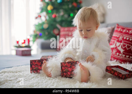 Curieux petit bébé garçon, vêtu de blanc handknitted dans l'ensemble, allongé sur la table jouant avec des cadeaux et heureusement smiling Banque D'Images