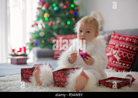 Curieux petit bébé garçon, vêtu de blanc handknitted dans l'ensemble, allongé sur la table jouant avec des cadeaux et heureusement smiling Banque D'Images