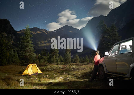 Camping avec une voiture jaune, tente à l'homme, nuit de lune avec phares avant en montagne Banque D'Images