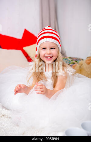 Petite fille dans un luxuriant robe blanche et un chapeau de Noël est assis sur le lit et de rires. Bonne Année et thème de Noël. Ambiance de fête. Banque D'Images