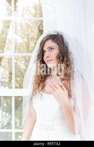 Closeup portrait de jeune femme, femme, mariée en robe de mariée, voile, le visage, les cheveux, collier de perles, debout, regardant par la fenêtre en verre, blanc Banque D'Images