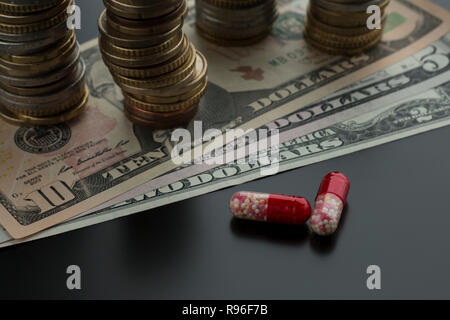 Deux comprimés ou capsules rouges contre des dollars et des piles de pièces de monnaie sur l'arrière-plan. Concept de traitement coûteux Banque D'Images