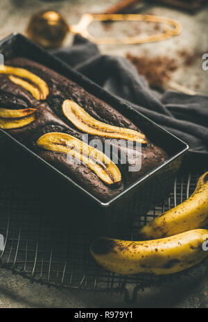 Du pain aux bananes et au chocolat noir dessert gâteau dans un moule sur une grille avec la cannelle, la poudre de cacao et de bananes fraîches sur le béton gris Banque D'Images