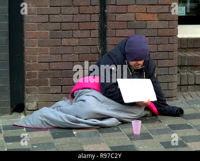 Sans-abri mendier dans rue avec mur de brique rose tasse se cacher face holding sign avec sac de couchage Banque D'Images