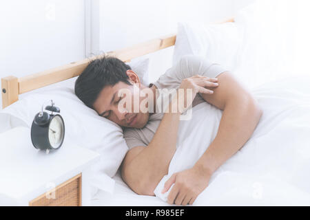 L'homme asiatique est en train de dormir sur le côté avec réveil lit blanc Banque D'Images