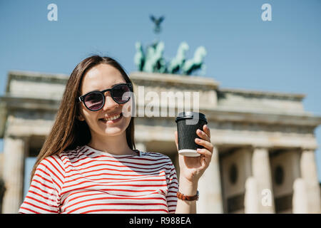 Portrait d'une belle jeune fille souriante positive qui tient dans sa main un café ou autre boisson sur l'arrière-plan de la porte de Brandebourg à Berlin en Allemagne. Banque D'Images