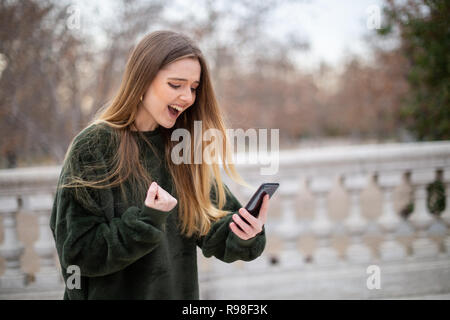 Jeune femme excitée criant et faisant des gestes avec la main en position debout dans le parc et à la recherche à smartphone moderne Banque D'Images