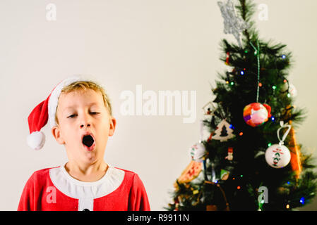 Blonde et beau garçon de 5 ans habillé en père Noël, ennuyé par l'arbre de Noël en attendant l'arrivée du vrai père, fond blanc Banque D'Images