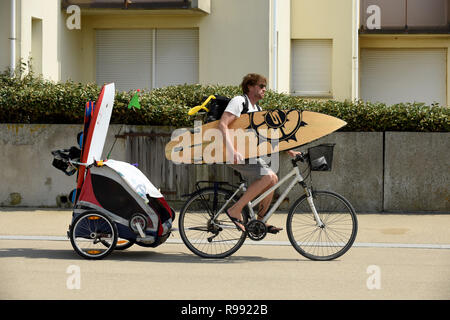 Surfer riding bike avec planche de surf et de remorquage remorque enfant à Wissant dans le nord de la France Banque D'Images