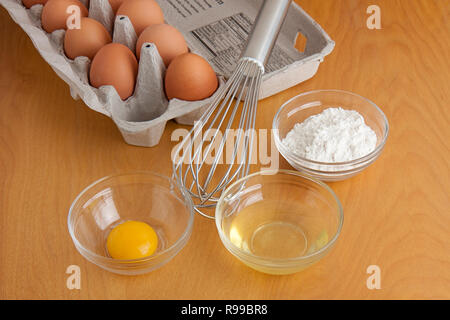 Séparés oeufs fêlés dans bols en verre, de la farine dans un bol en verre, un fouet d'argent, et la boîte d'oeufs tous les sur une planche à découper. Banque D'Images