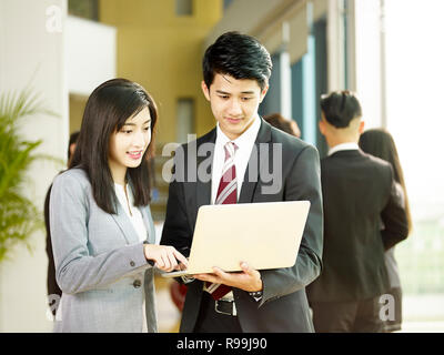 Young Asian businessman and businesswoman working together in office using laptop computer, collègues parler en arrière-plan. Banque D'Images