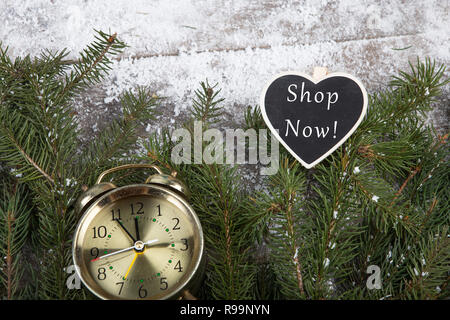 Réveil, tableau noir en forme de coeur avec l'inscription 'Shop maintenant !' et le sapin arbre branche sur un fond de bois couverte de neige Banque D'Images