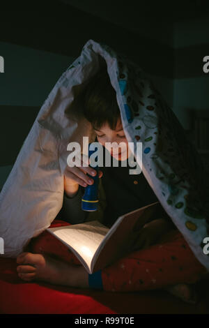 Garçon lisant un livre avec une lampe de poche sous la couette Banque D'Images