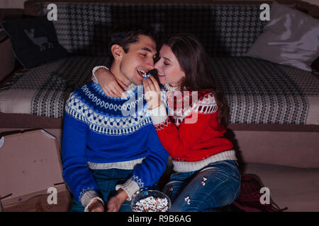 Jeune couple watching movie eating popcorn at home la nuit. Woman feeding her boyfriend Banque D'Images