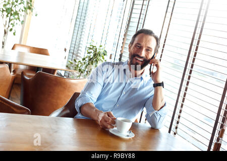 Commerçant homme mûr assis à table à café à la tenue de jour tasse de café chaud à parler sur smartphone rire joyeux Banque D'Images