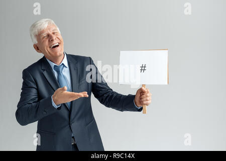 Senior business man studio isolés sur mur gris holding white board montrant dièse à la joie de rire de l'appareil photo Banque D'Images