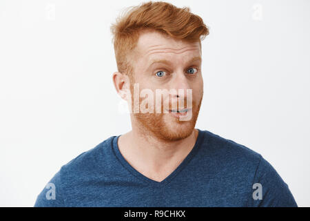 Portrait de l'intrigué beau mâle avec gingembre en soie t-shirt bleu tourner en caméra avec impressionné et curieux, à l'écoute de l'expression histoire intéressante avec précaution sur fond gris Banque D'Images